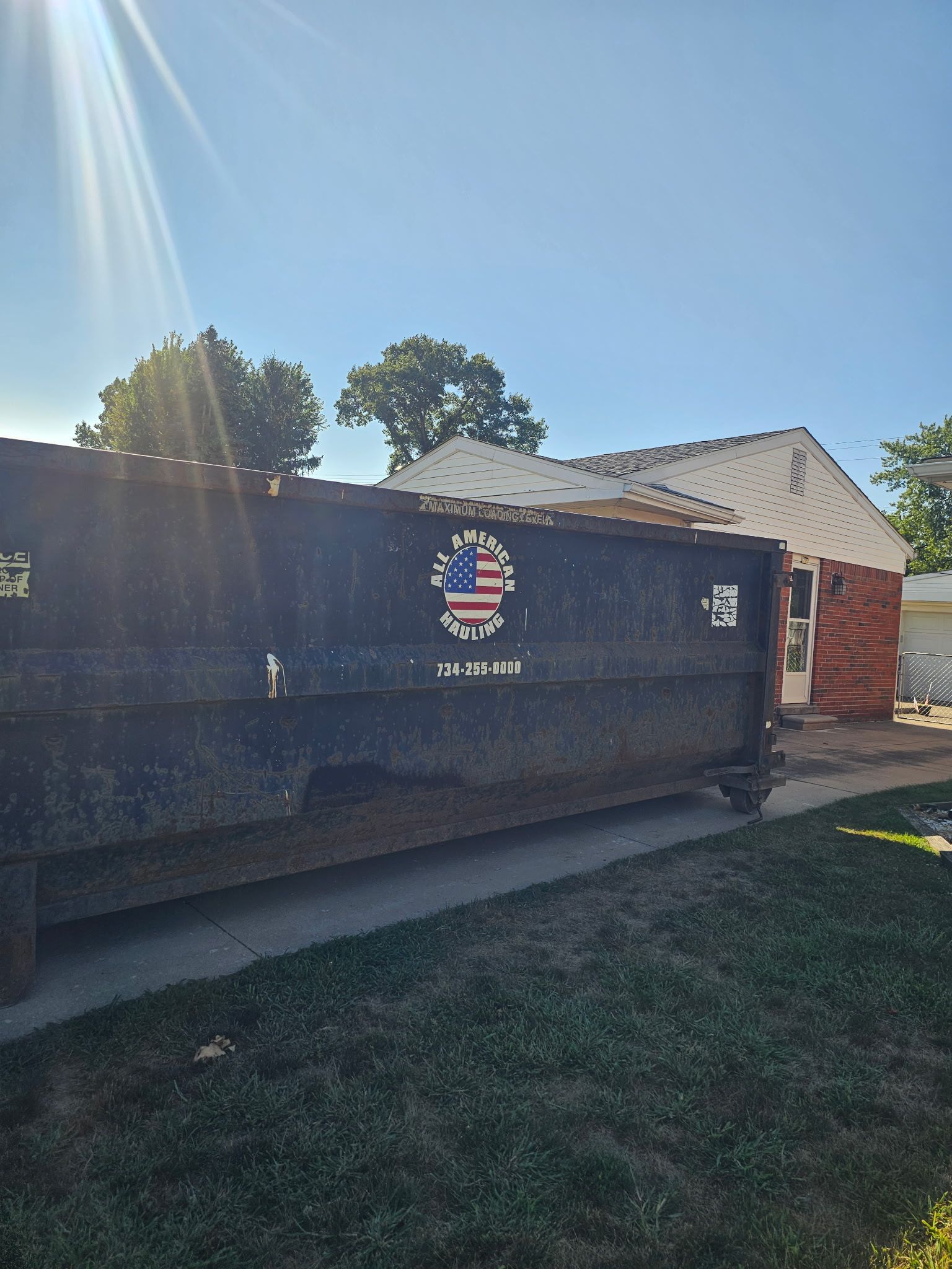 Large black dumpster in a sunny yard beside a house, with a red-sided building in the background.