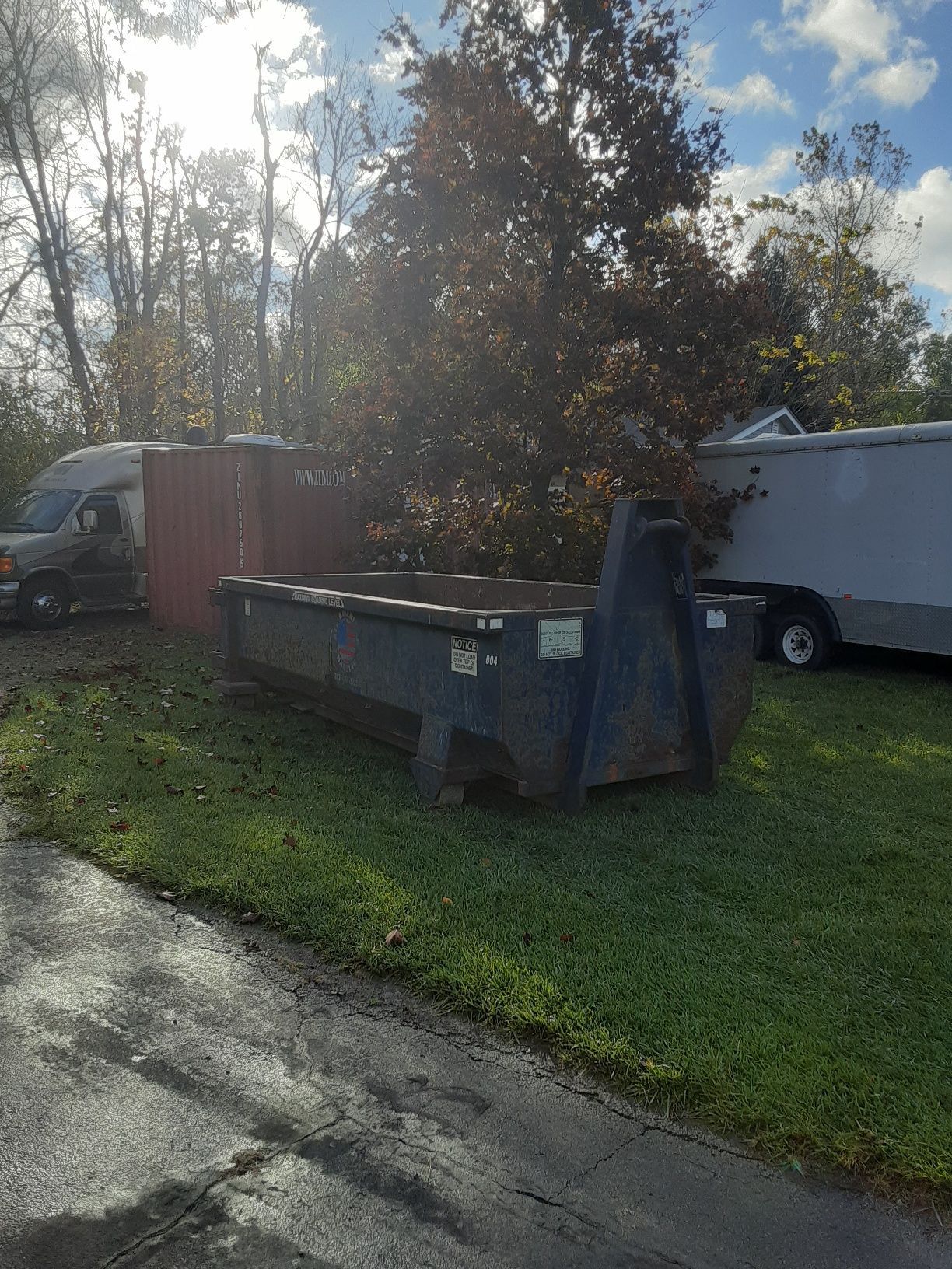 A dark blue roll-off dumpster sits on a grassy lawn near a parked truck and trailer on a sunny day.