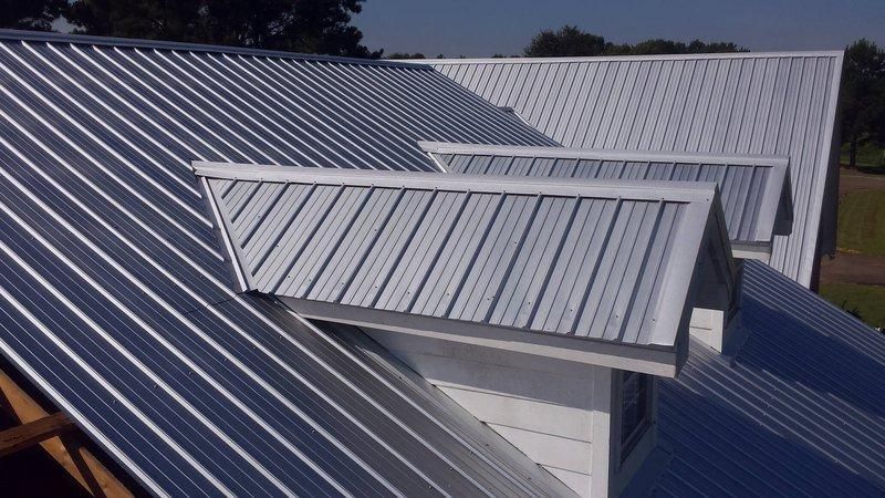 Metal-roof dormer on a gray roof under sunlight, with nearby trees in the background