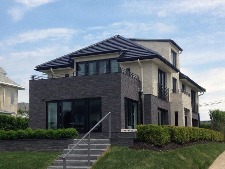 Modern two-story brick house with gray roof, front steps, and trimmed hedges under a blue sky