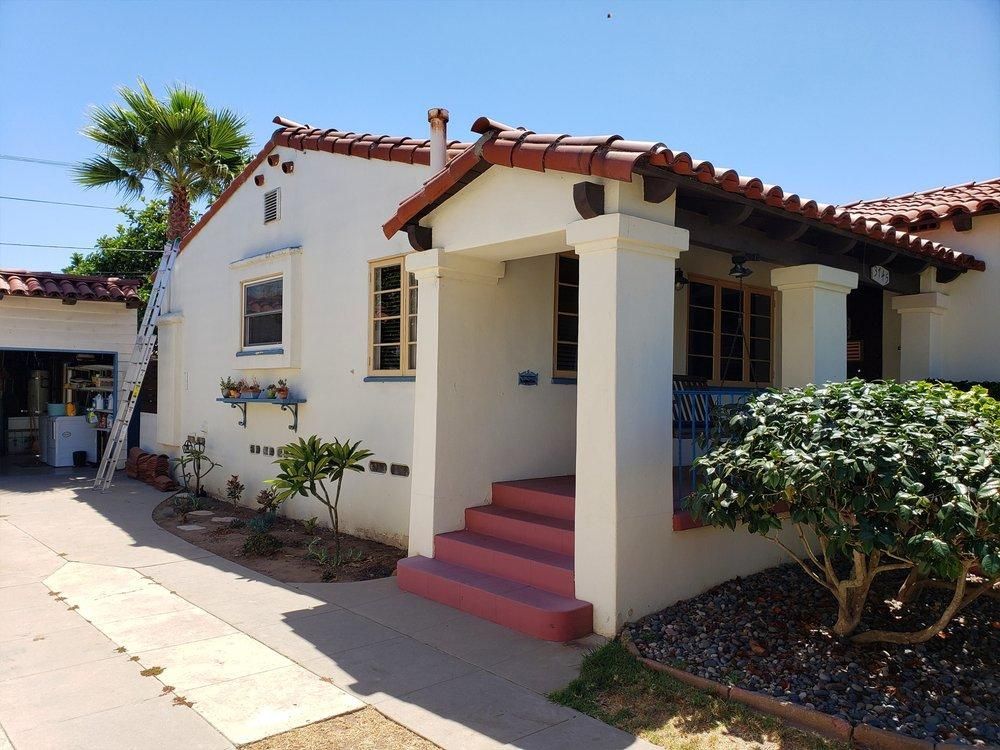 Small white stucco house with red steps, terracotta roof, and front porch under a blue sky