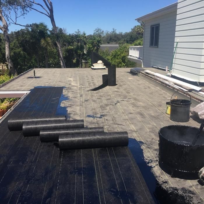 Roof under repair with rolled black roofing material on a flat rooftop beside a house.