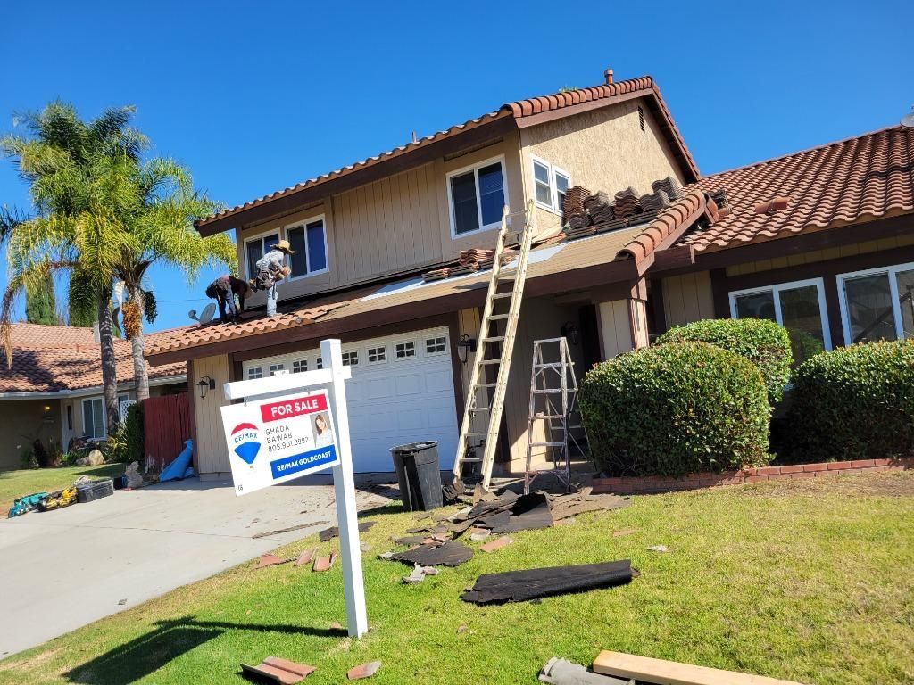 Two workers on a ladder repairing a two-story house roof beside a real estate sign.