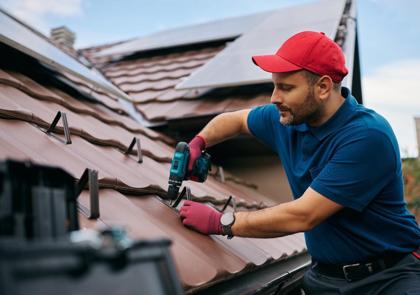 Worker in red cap installing a roof vent on brown shingles with a power drill