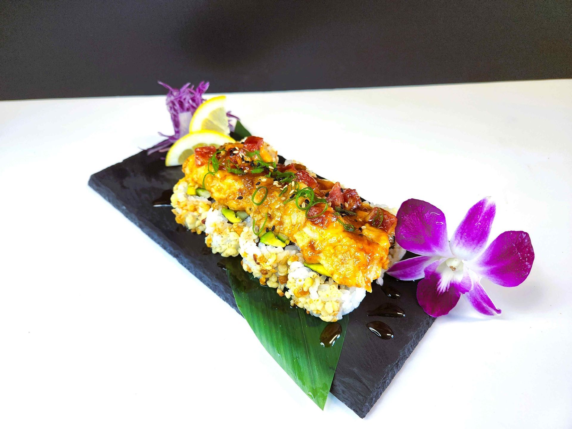 A sushi roll with a purple flower and a lemon on a black plate.