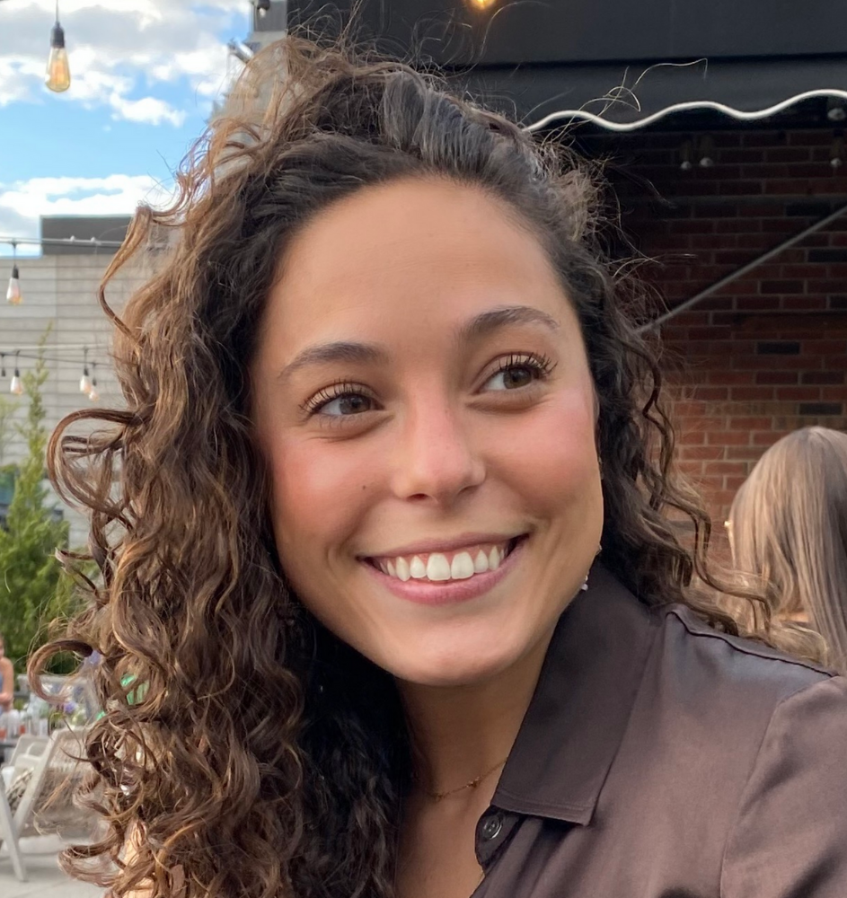 Woman with curly brown hair smiles outdoors, wearing a brown shirt.