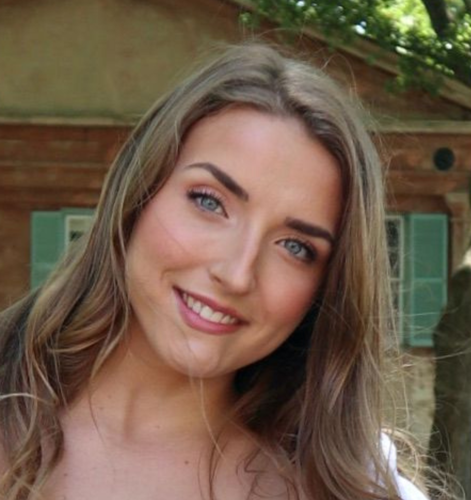 Woman with long blonde hair smiles outdoors near a building with green shutters.