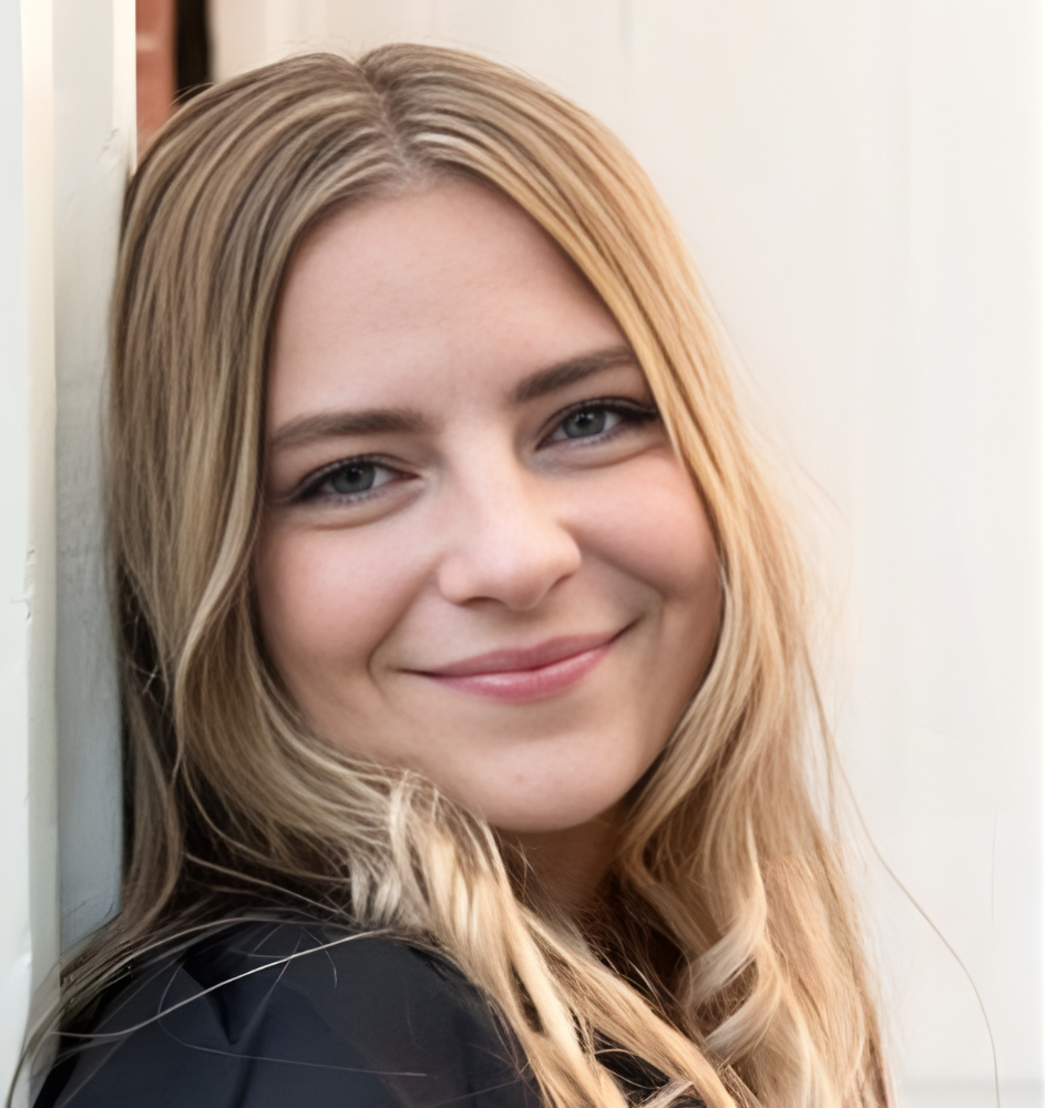 Woman with blonde hair smiles, leaning against a white wall outdoors.