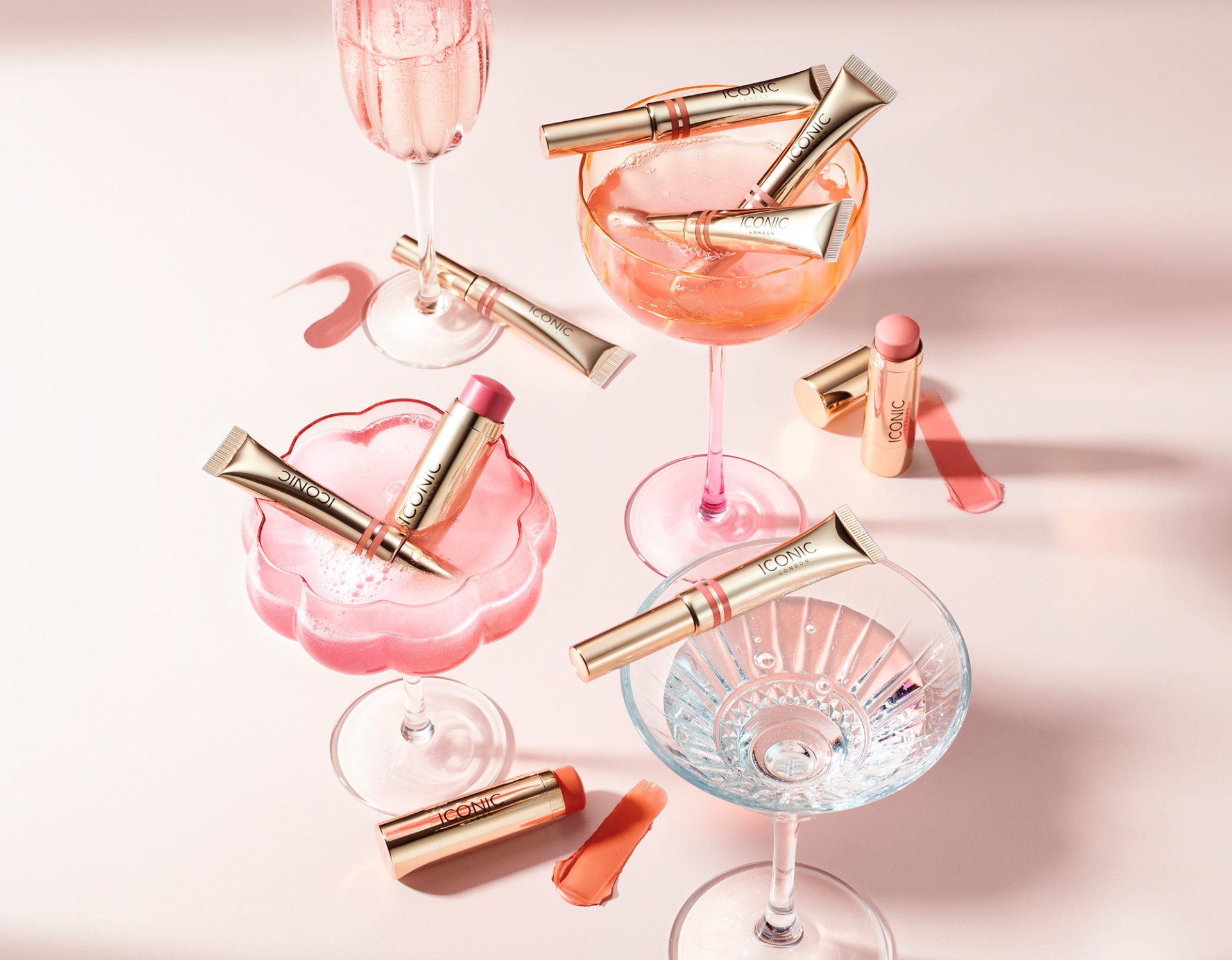 Makeup products arranged in and around pink and clear glassware on a pink surface.