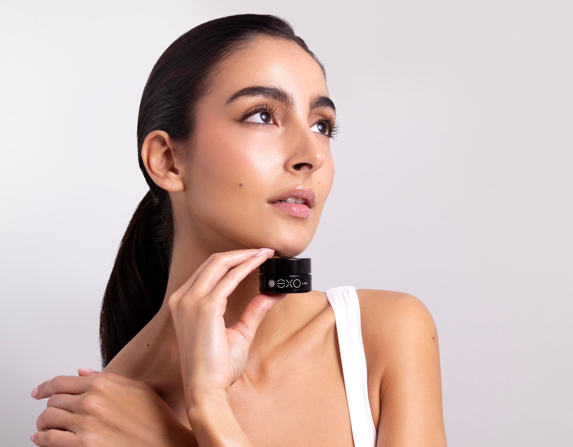 Woman holding a small black jar, looking up with a soft expression. White top, light background.