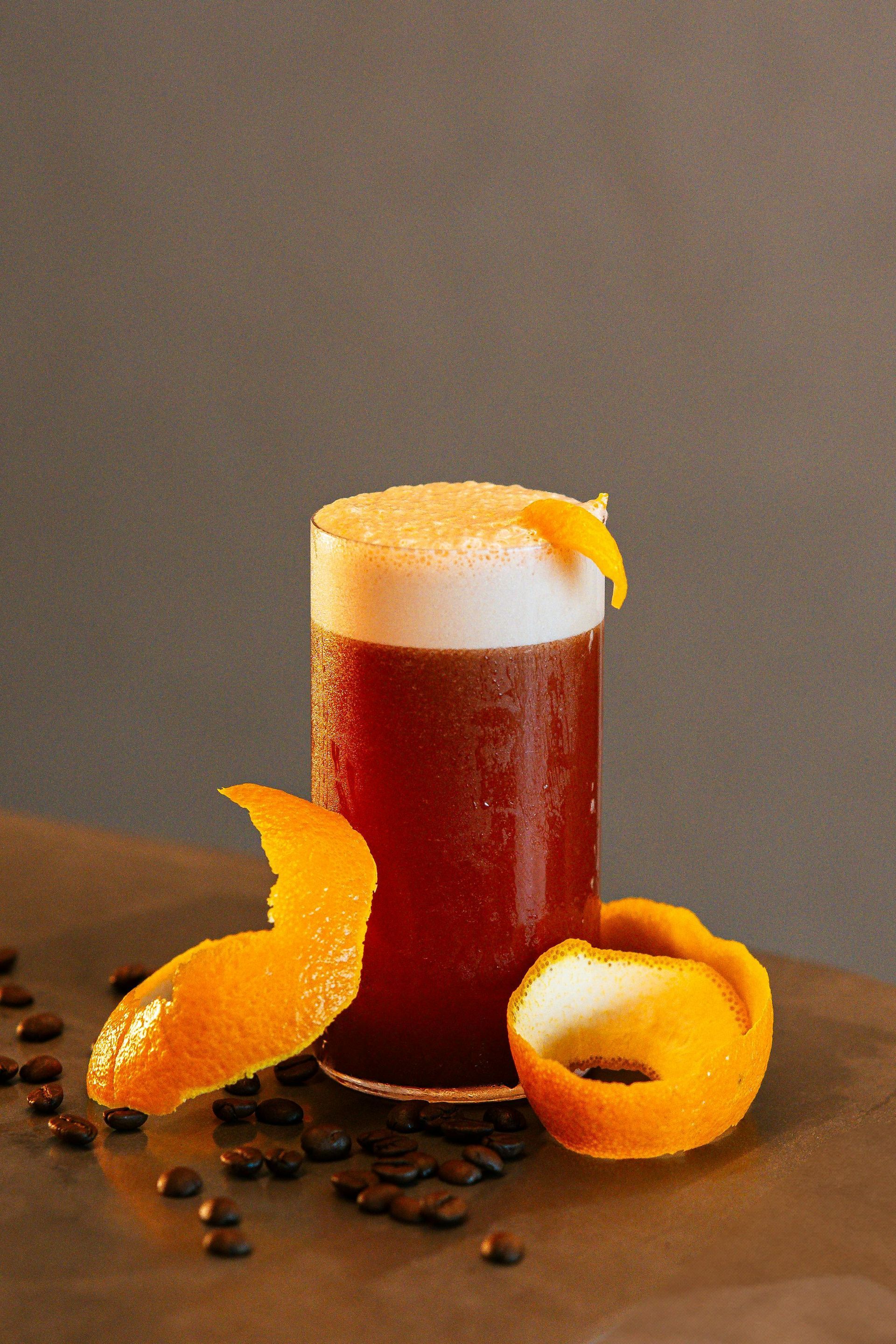 Coffee cocktail in tall glass, topped with foam, orange peel garnish, coffee beans, on gray surface.