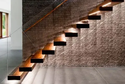 A wooden staircase with a glass railing is against a brick wall.