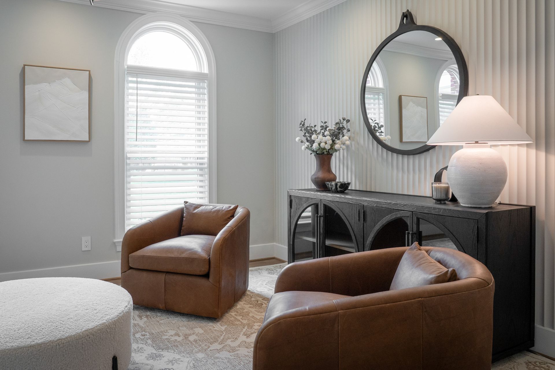 A living room with two chairs , an ottoman , a mirror and a window.