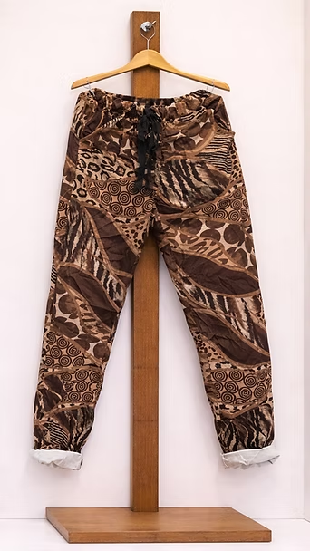 Pantalones marrones estampados colgados en un soporte de madera sobre un fondo blanco.