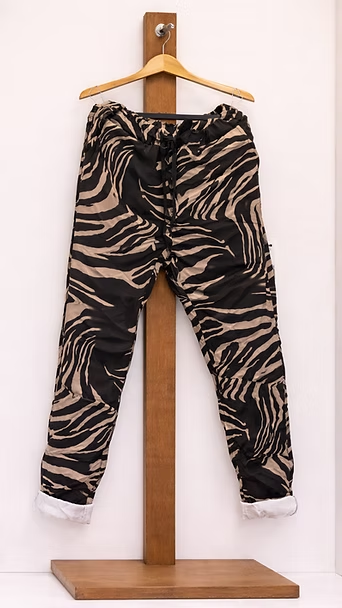 Pantalones con estampado de cebra en negro y marrón colgando de un soporte de madera sobre un fondo claro.