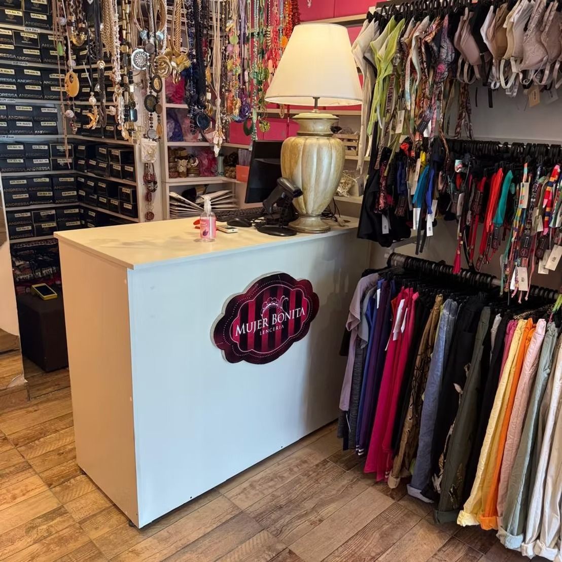 Mostrador de una pequeña boutique con lámpara, expositores de joyería y percheros con accesorios y ropa de colores.