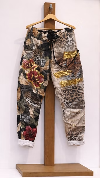 Pantalones estampados colgados en un perchero de madera, con estampados florales y abstractos en negro, beige y rojo.