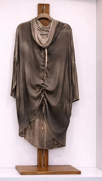 Vestido túnica gris de manga larga con drapeado, colgado de un soporte de madera sobre un fondo blanco.