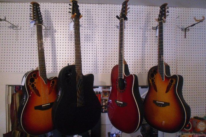 Four acoustic guitars hanging on a pegboard wall, in orange, black, red, and sunburst finishes.