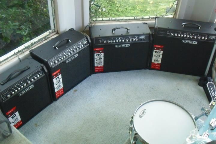 Small drum kit surrounded by black guitar amplifiers in a carpeted room by large windows