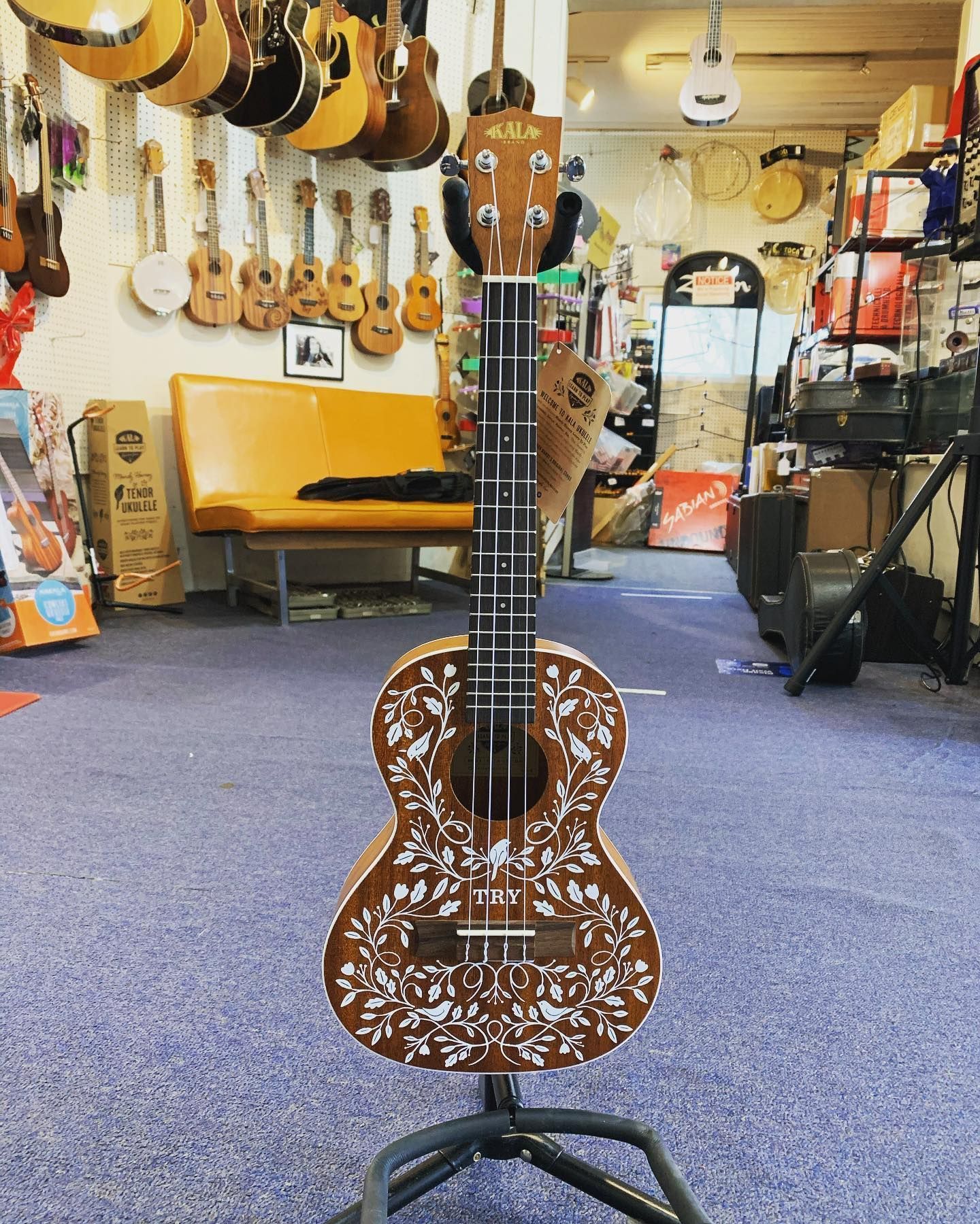 Brown floral-engraved ukulele on a stand in a music shop, with guitars hanging in the background