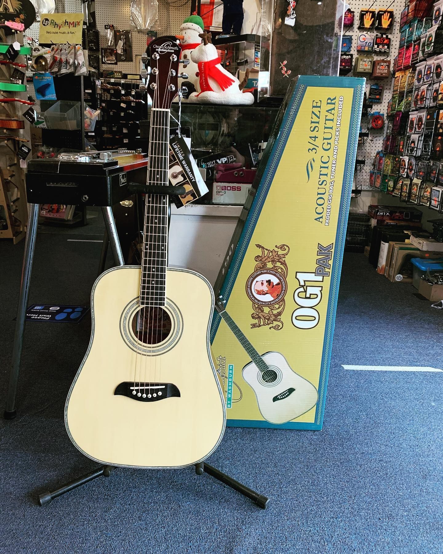 Acoustic guitar on a stand in a music shop, beside a boxed OGIG guitar set.