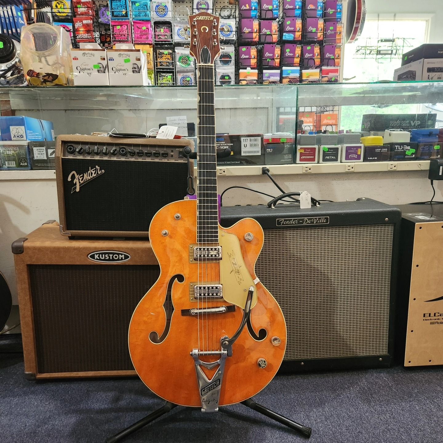Orange hollow-body electric guitar on a stand in a music shop, with amplifiers and shelves behind.