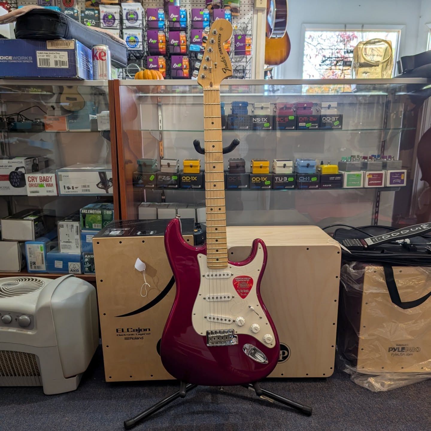Red electric guitar on a stand in a music shop, with shelves of gear and boxes behind it.