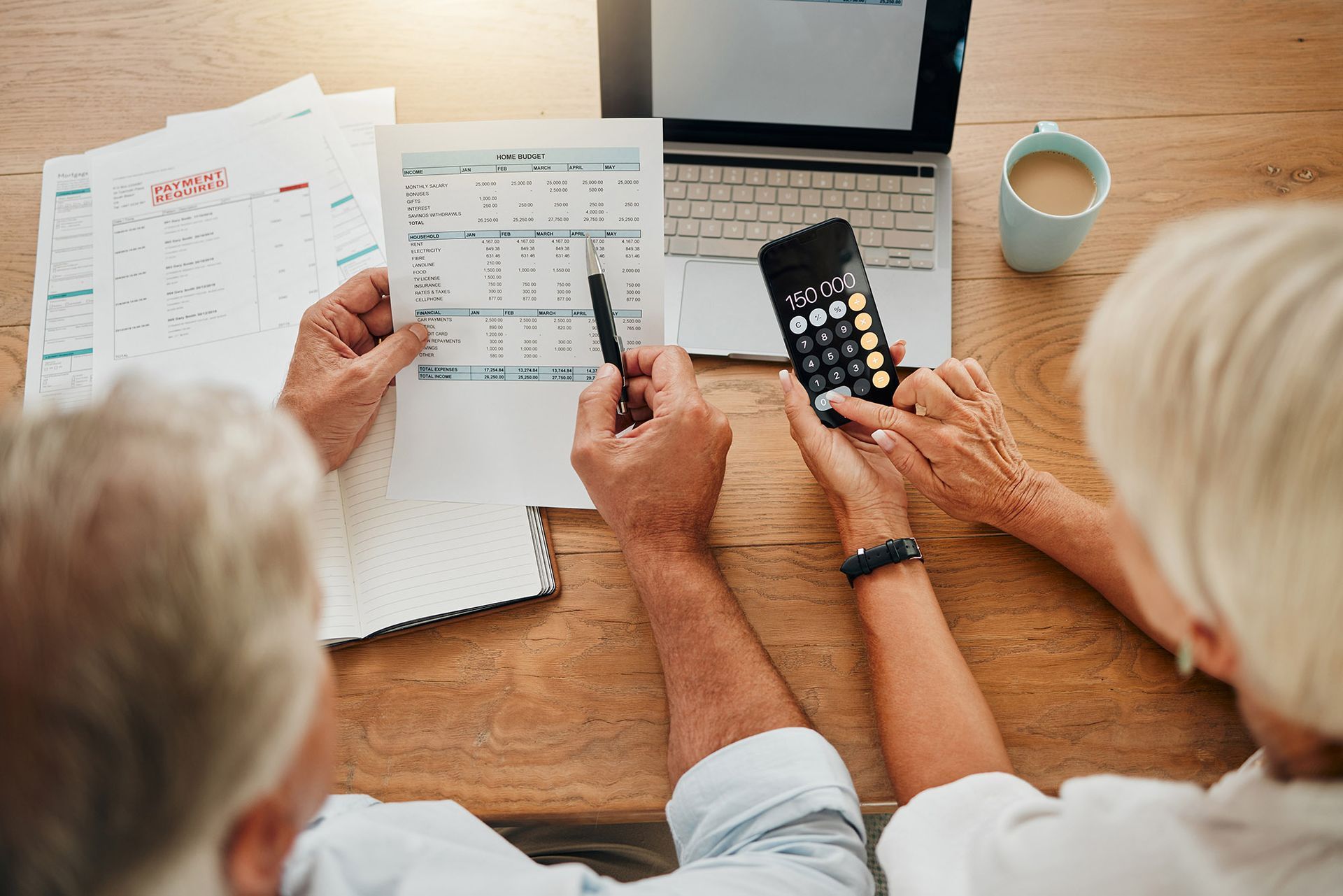 Couple reviewing financial documents, using a calculator and laptop, with coffee cup on a wooden table.