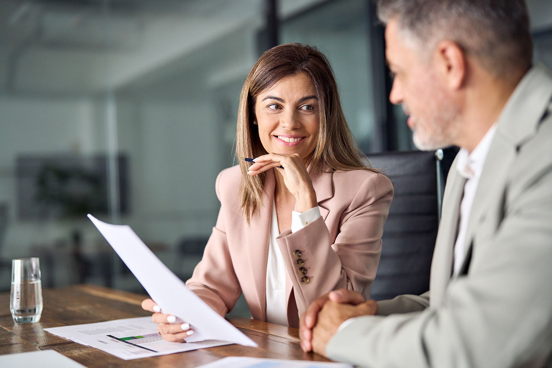 Woman in pink blazer smiles at document, discussing with man at a table.