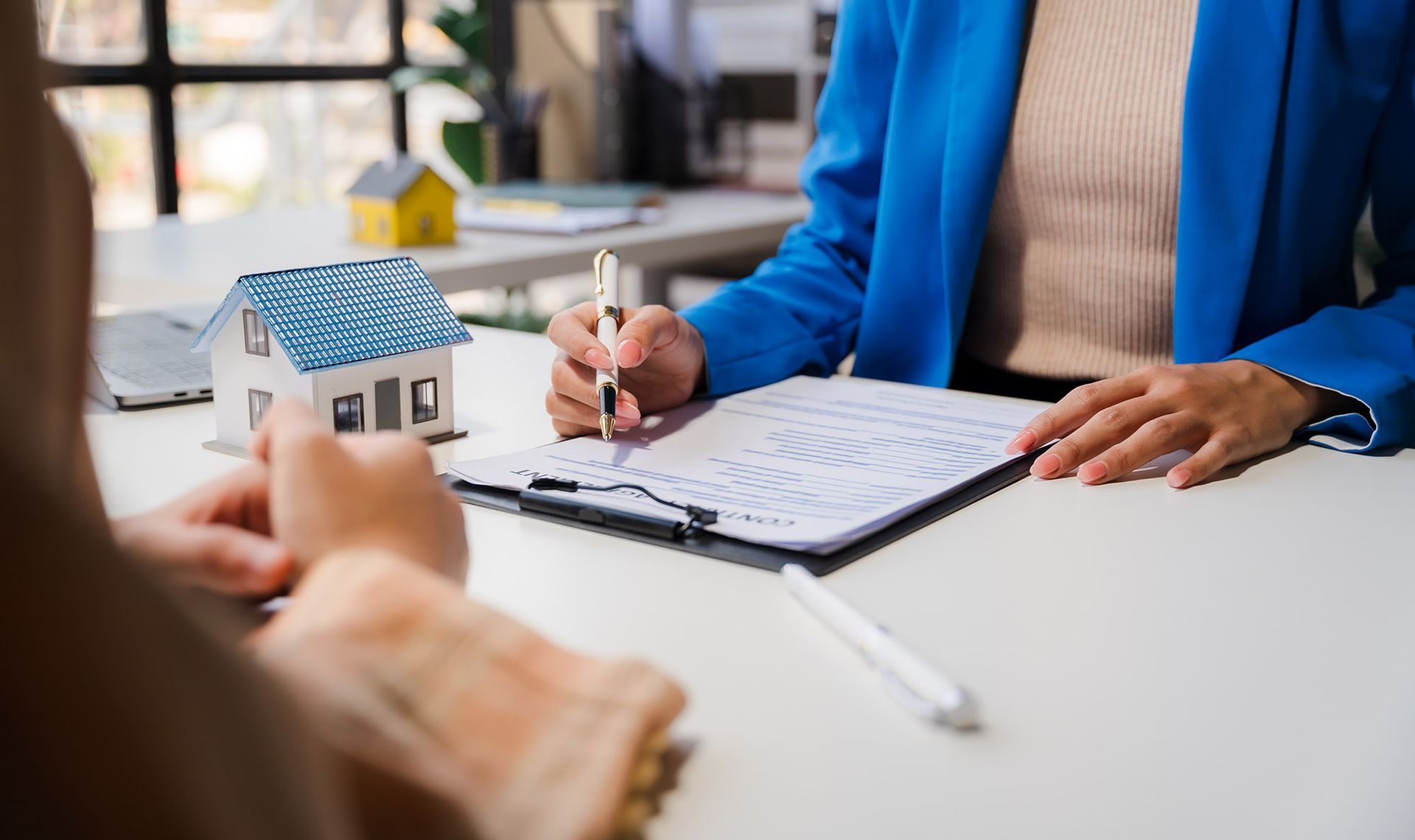 Person signing paperwork with a pen, a miniature house, and a phone on the table.