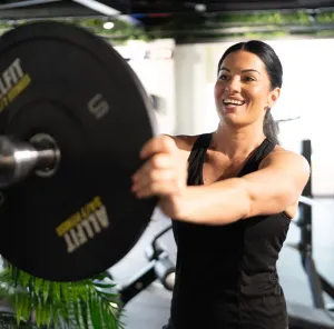 Woman lifting a barbell plate in a gym, smiling in a black workout top.