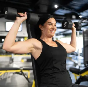 Woman lifting dumbbells on a bench in a gym, wearing a black tank top and smiling