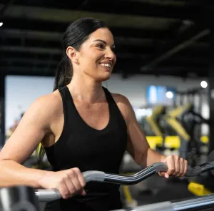 Woman using a gym machine, smiling in a black tank top, with blurred equipment in the background