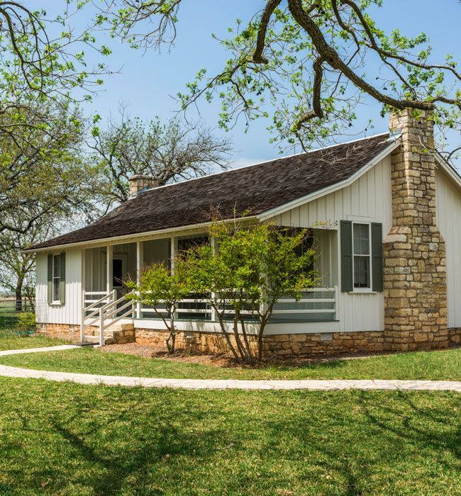 White cottage with stone chimney and porch in a grassy yard under trees