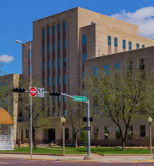 Downtown street corner with a tan office building, trees, and road signs under a blue sky