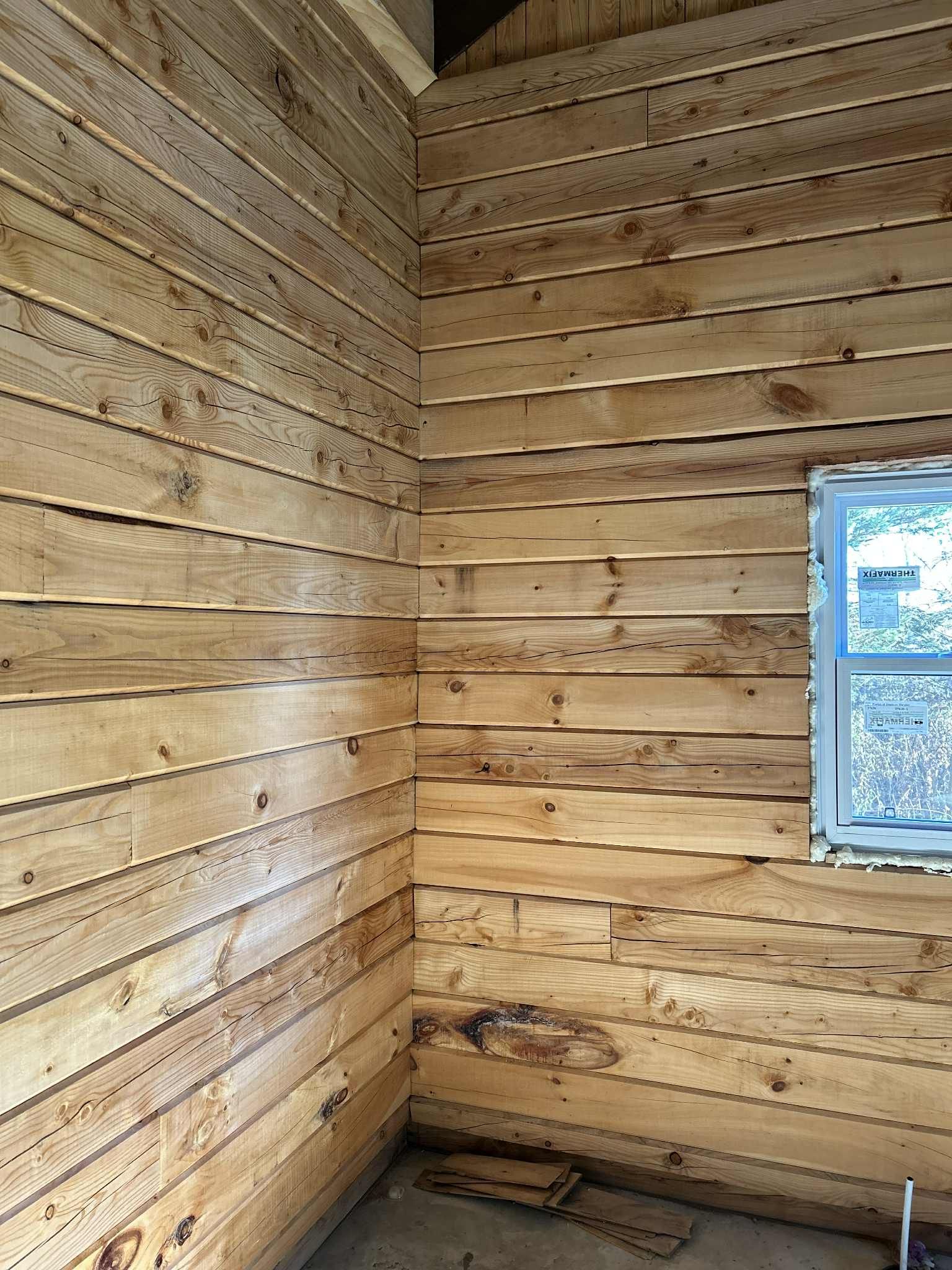 Murs intérieurs lambrissés en bois dans une pièce, avec une fenêtre et des trous de clous apparents.