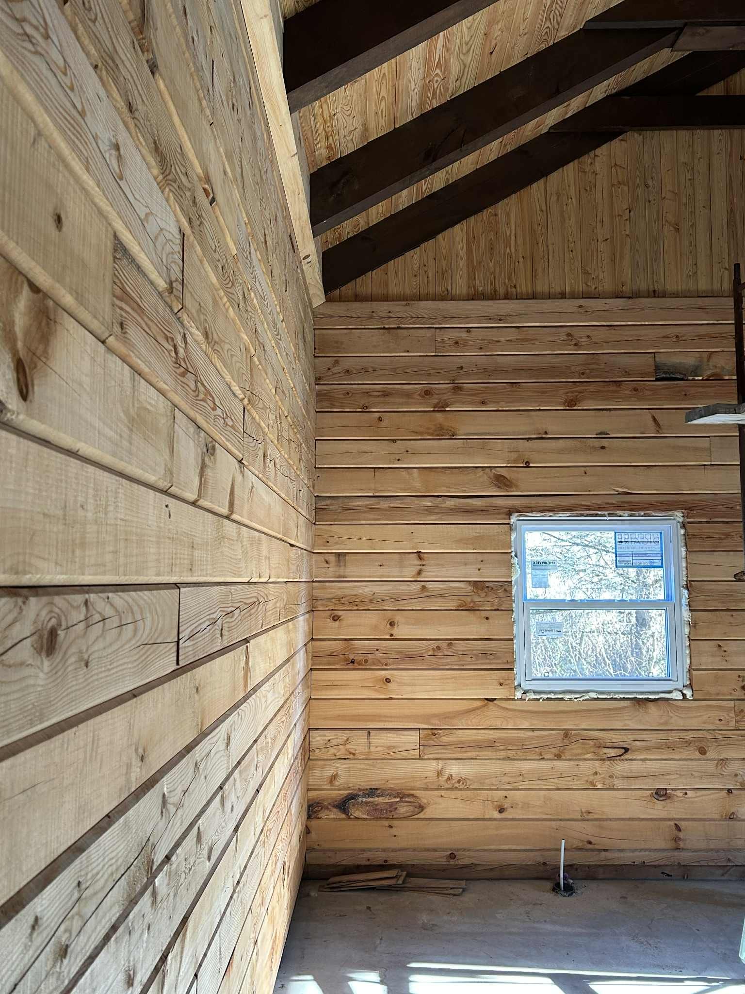 Intérieur d'une structure en bois en construction, avec des planches horizontales et verticales. Une petite fenêtre est visible.