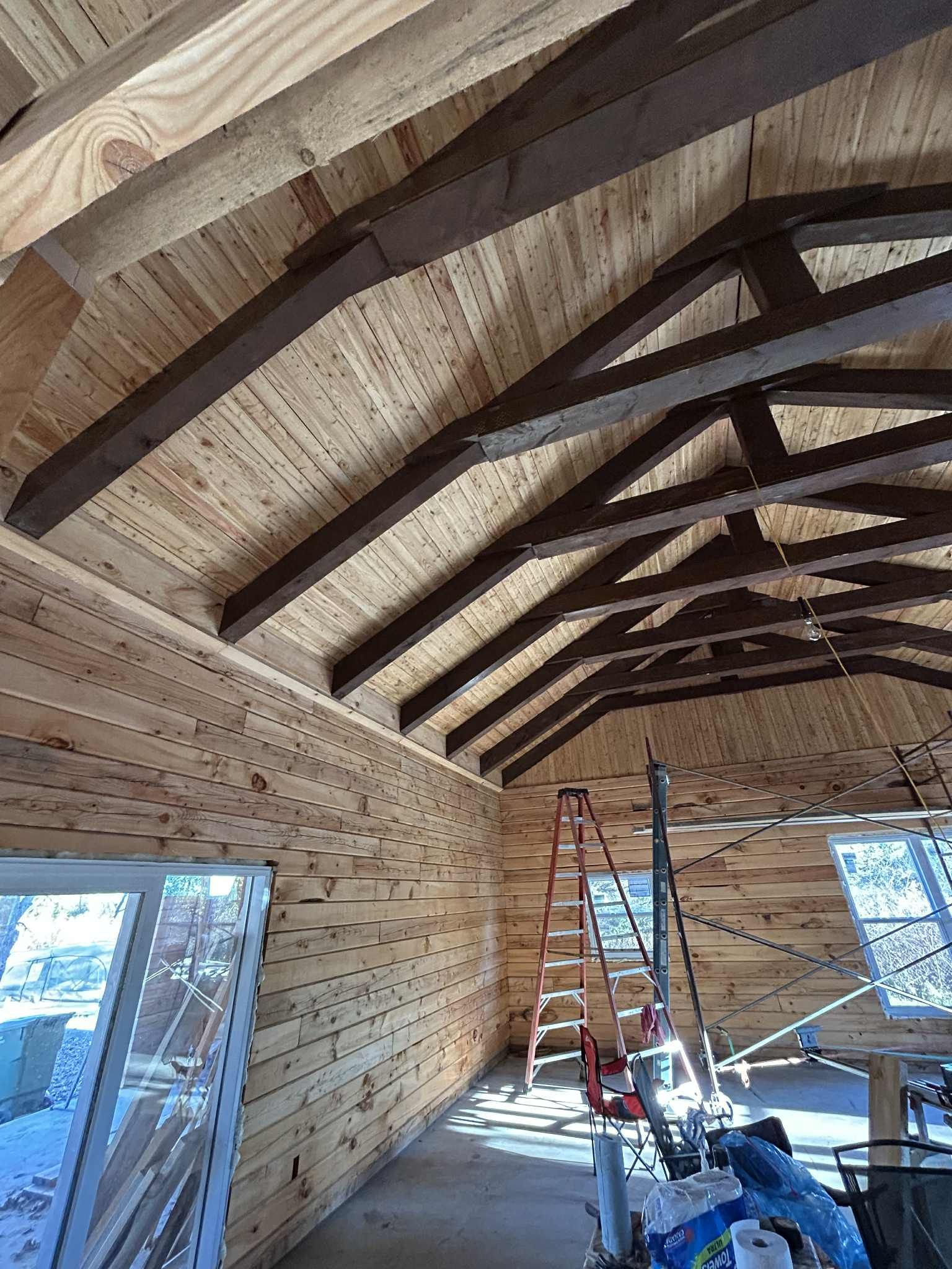 Intérieur d'un bâtiment en bois en construction. Des poutres brunes soutiennent un plafond en bois. Une échelle rouge se trouve à l'intérieur.