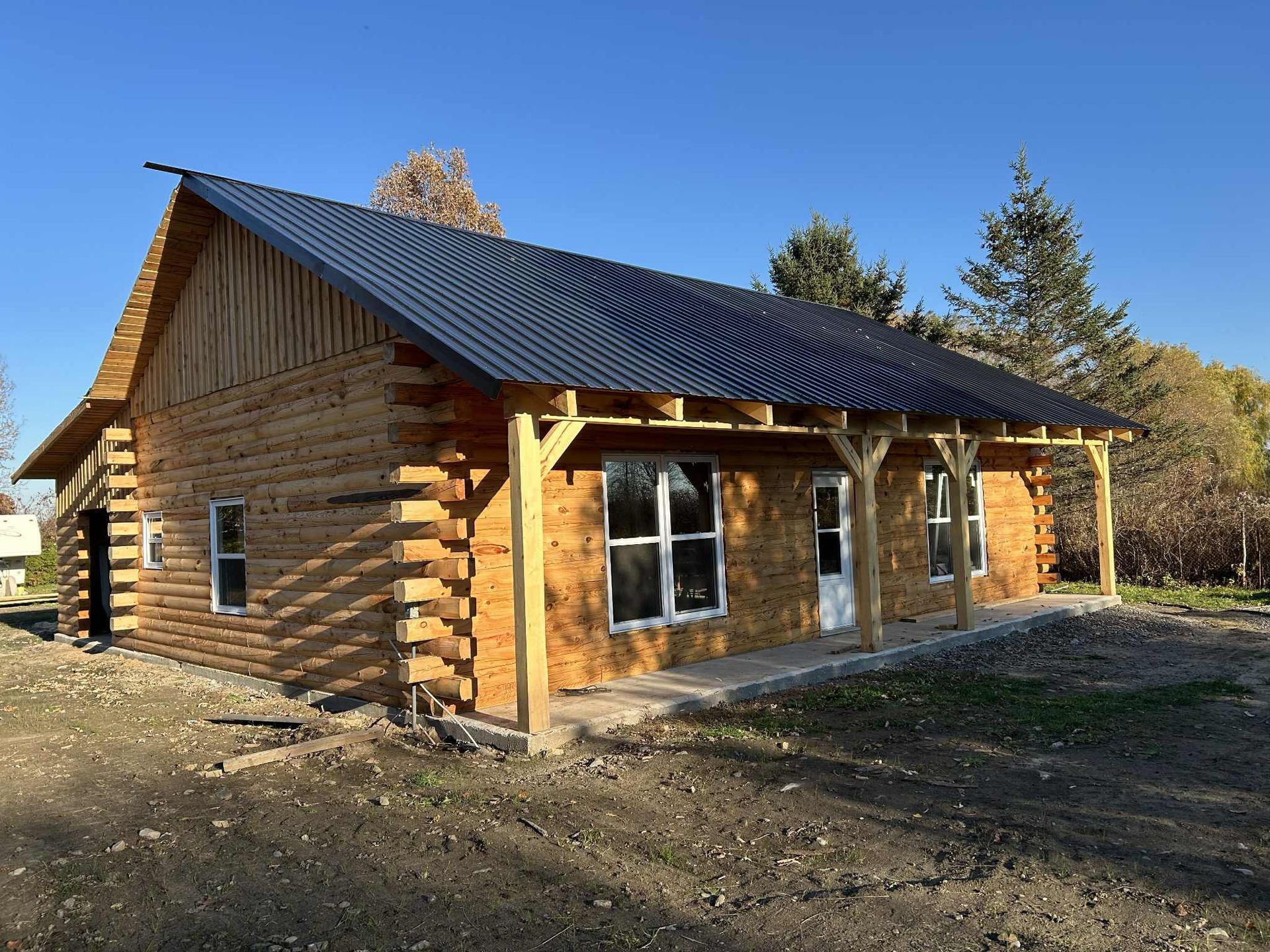 Cabane en rondins en construction, avec un toit en tôle ondulée noire et un porche.