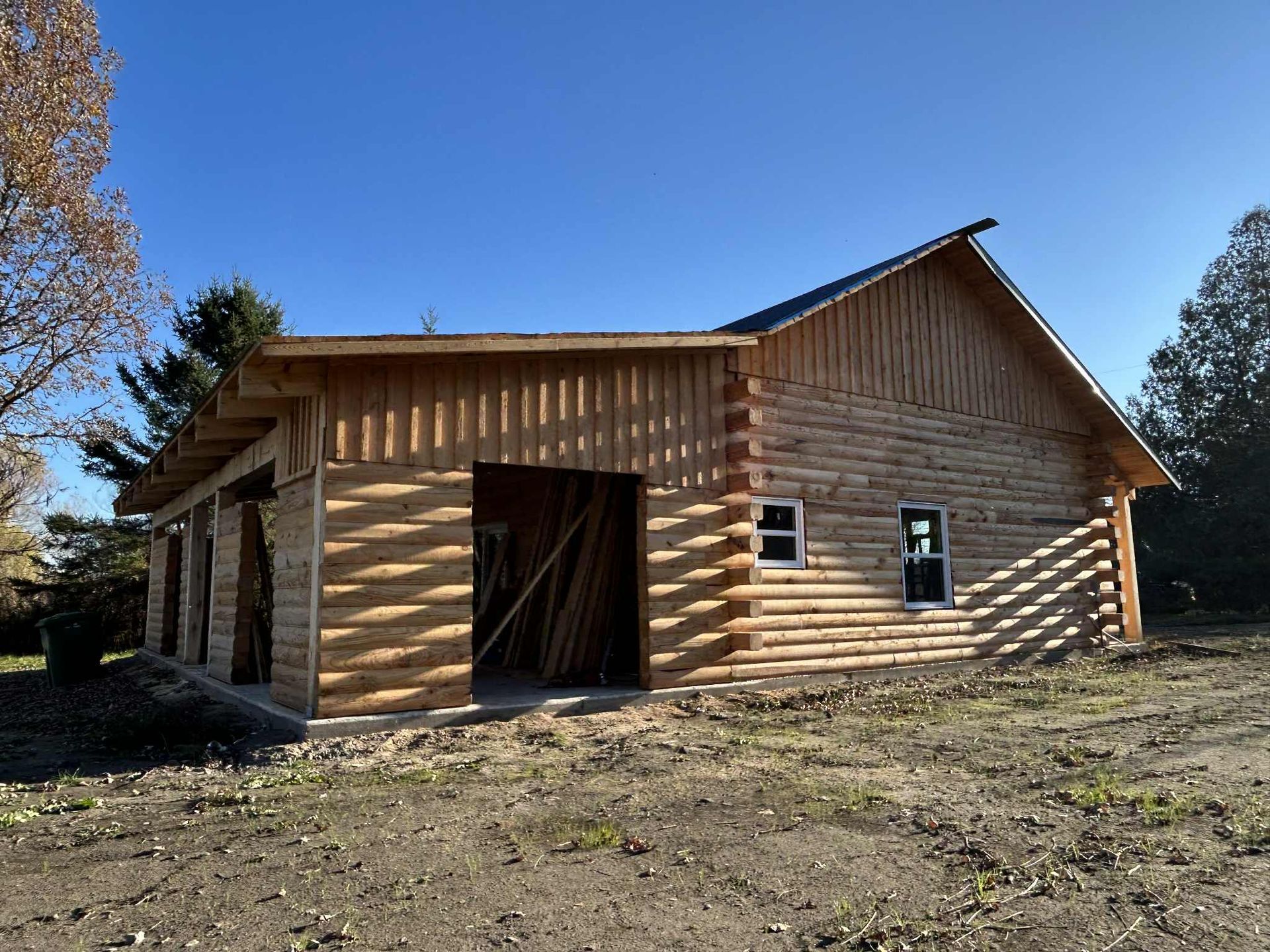 Chalet en rondins avec garage ouvert sur un côté et toit à pignon, en construction, à l'extérieur par une journée ensoleillée.