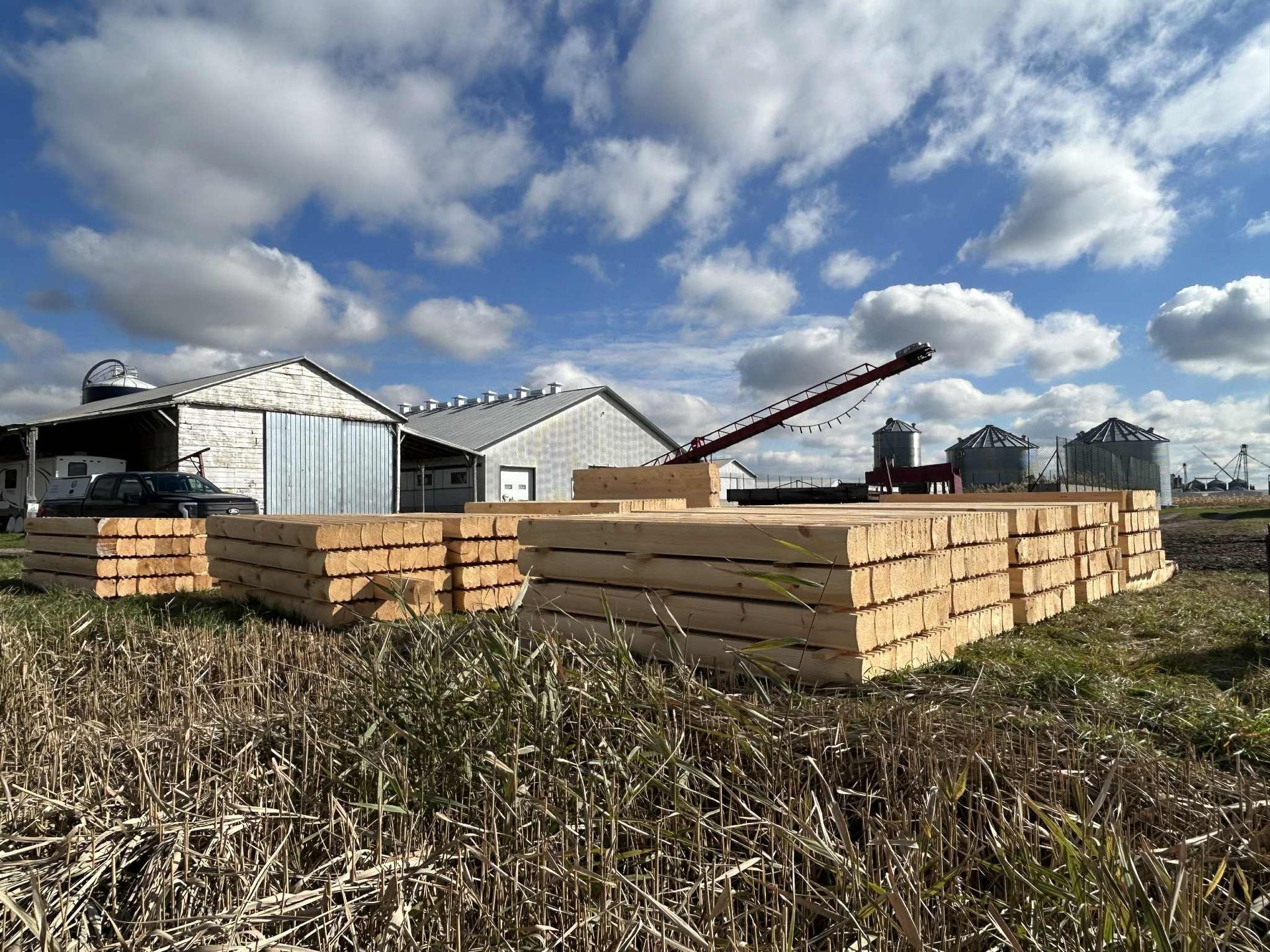 Des piles de bois dans un champ, avec des bâtiments agricoles et des silos à grains, sous un ciel bleu nuageux.