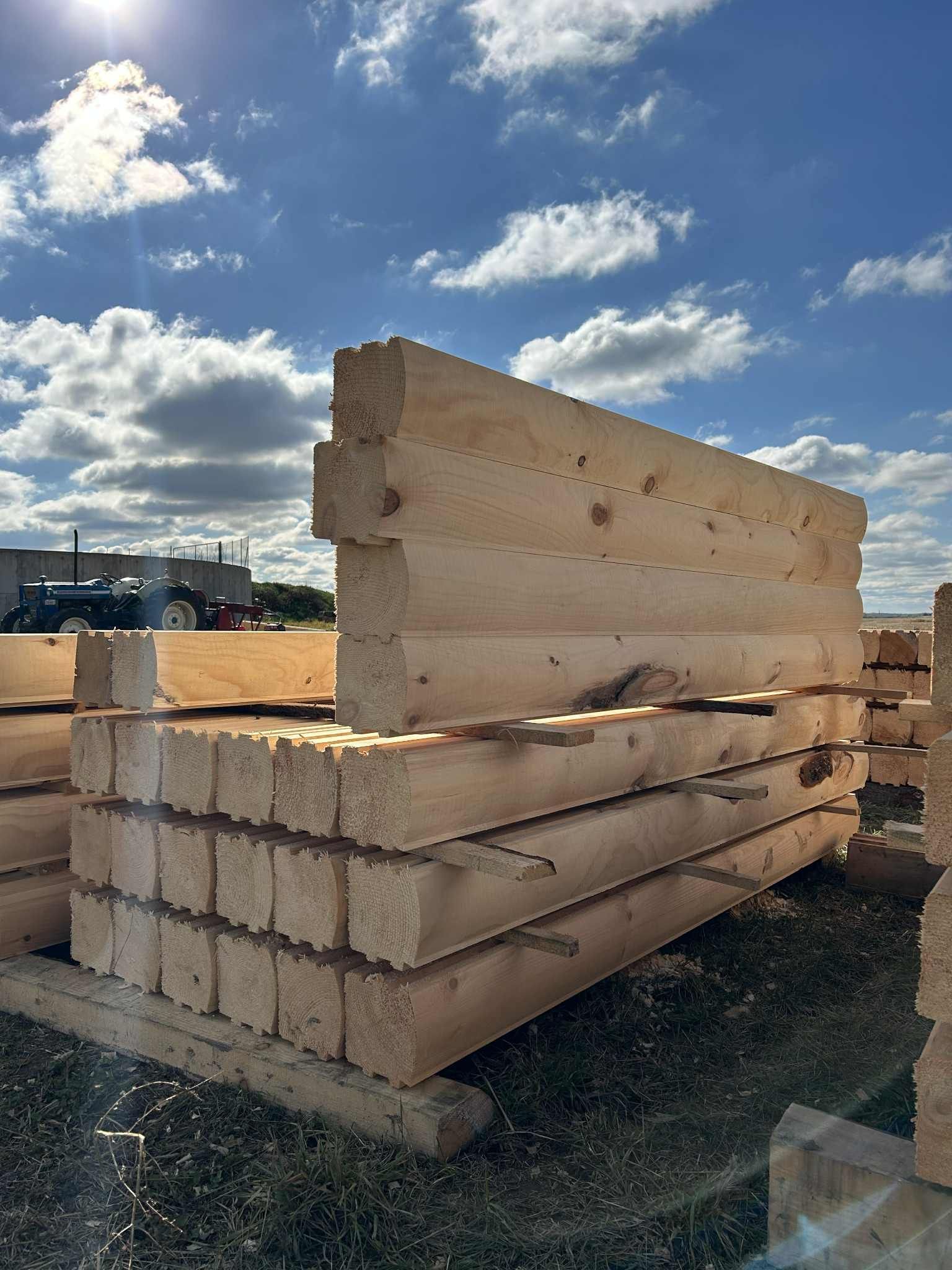 Empilement de poutres en bois inachevées à l'extérieur, sous un ciel bleu parsemé de nuages épars.