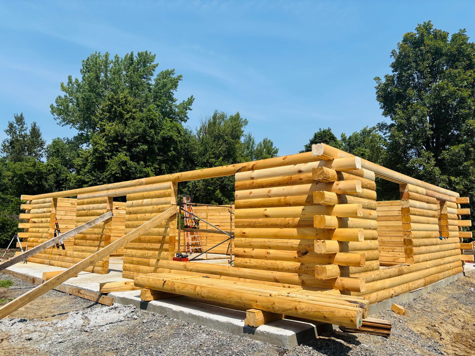 Cabane en rondins en construction, avec des rondins de bois empilés sur des fondations en béton, ciel bleu clair en arrière-plan.