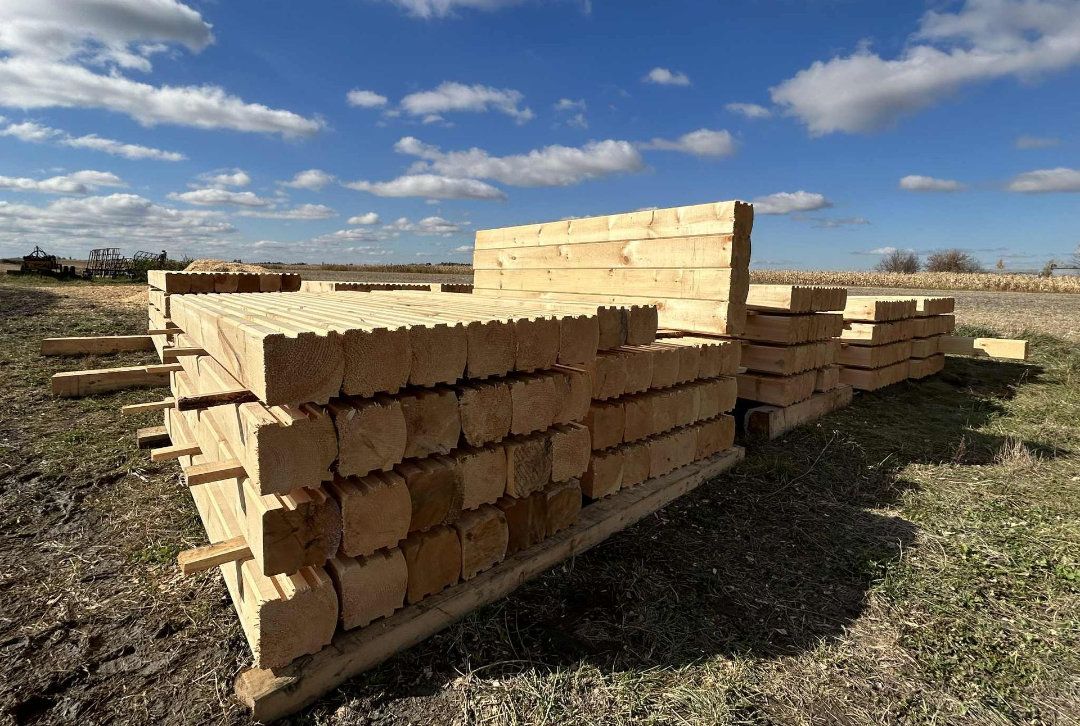 Des piles de poutres de bois à section carrée sur une palette en bois, à l'extérieur, sous un ciel bleu parsemé de nuages ​​épars.