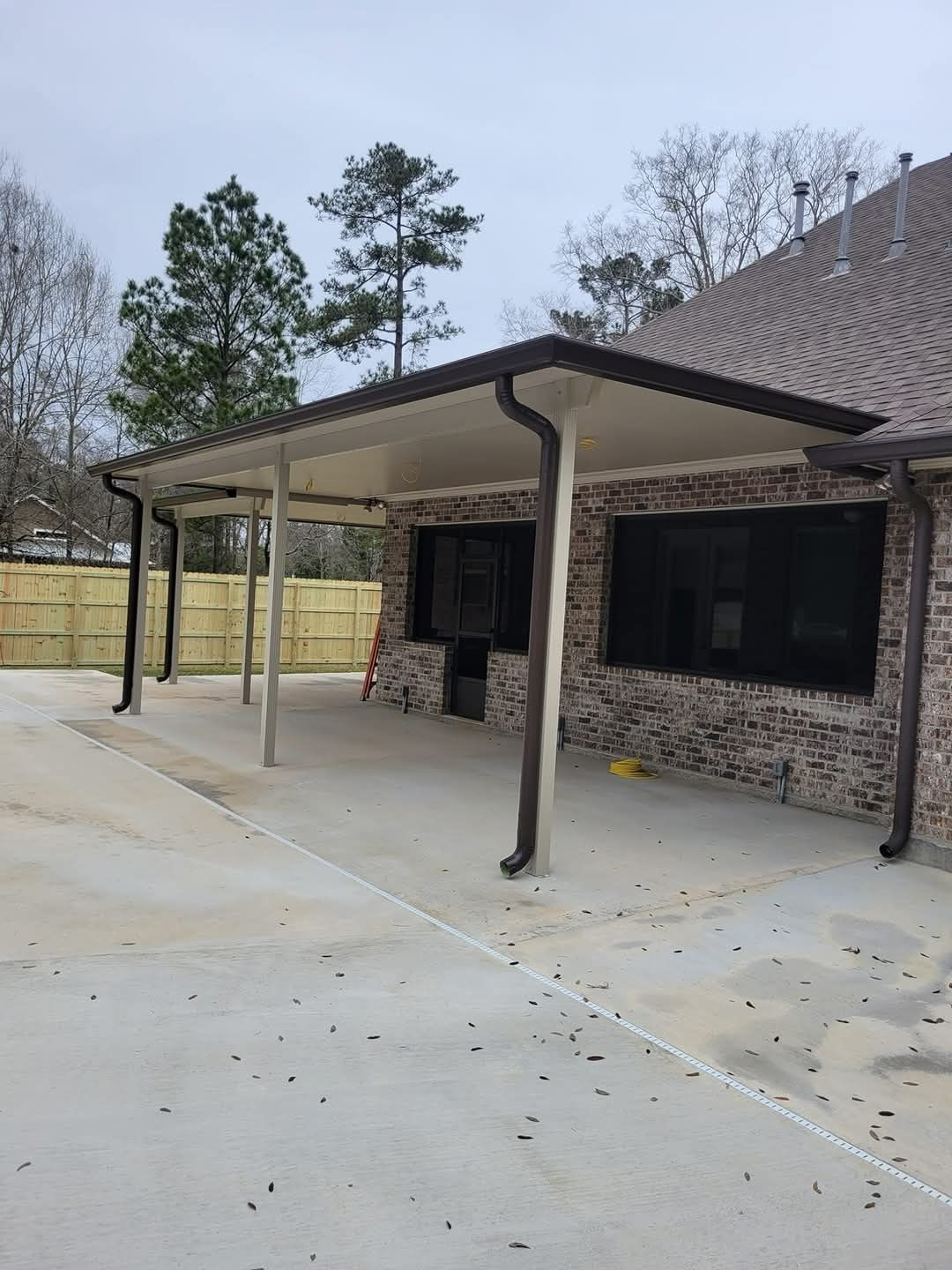 Covered patio beside a brick house with concrete driveway and leafless trees nearby