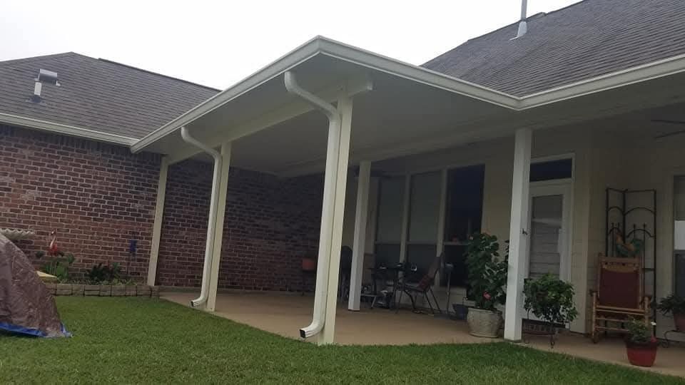Covered patio beside a brick house, with white columns, lawn, and potted plants.