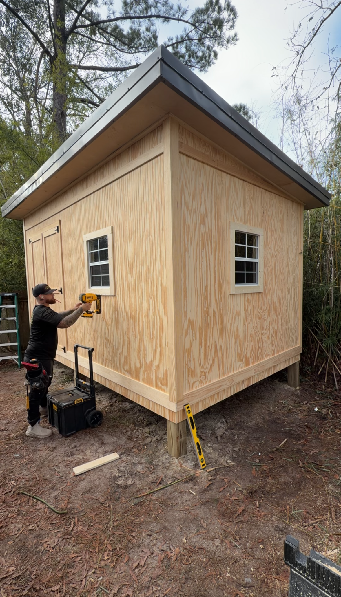 Person using a drill beside a small unfinished wooden cabin with a gray roof in a wooded area
