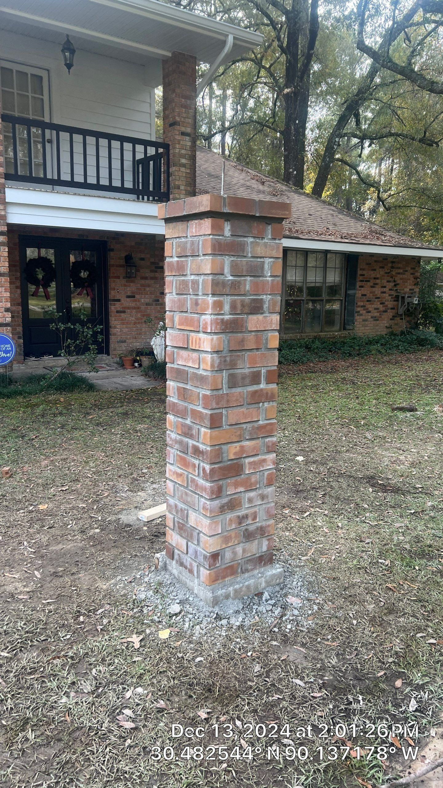 Brick column on a front lawn in front of a house, with a second-story balcony and trees in the background.