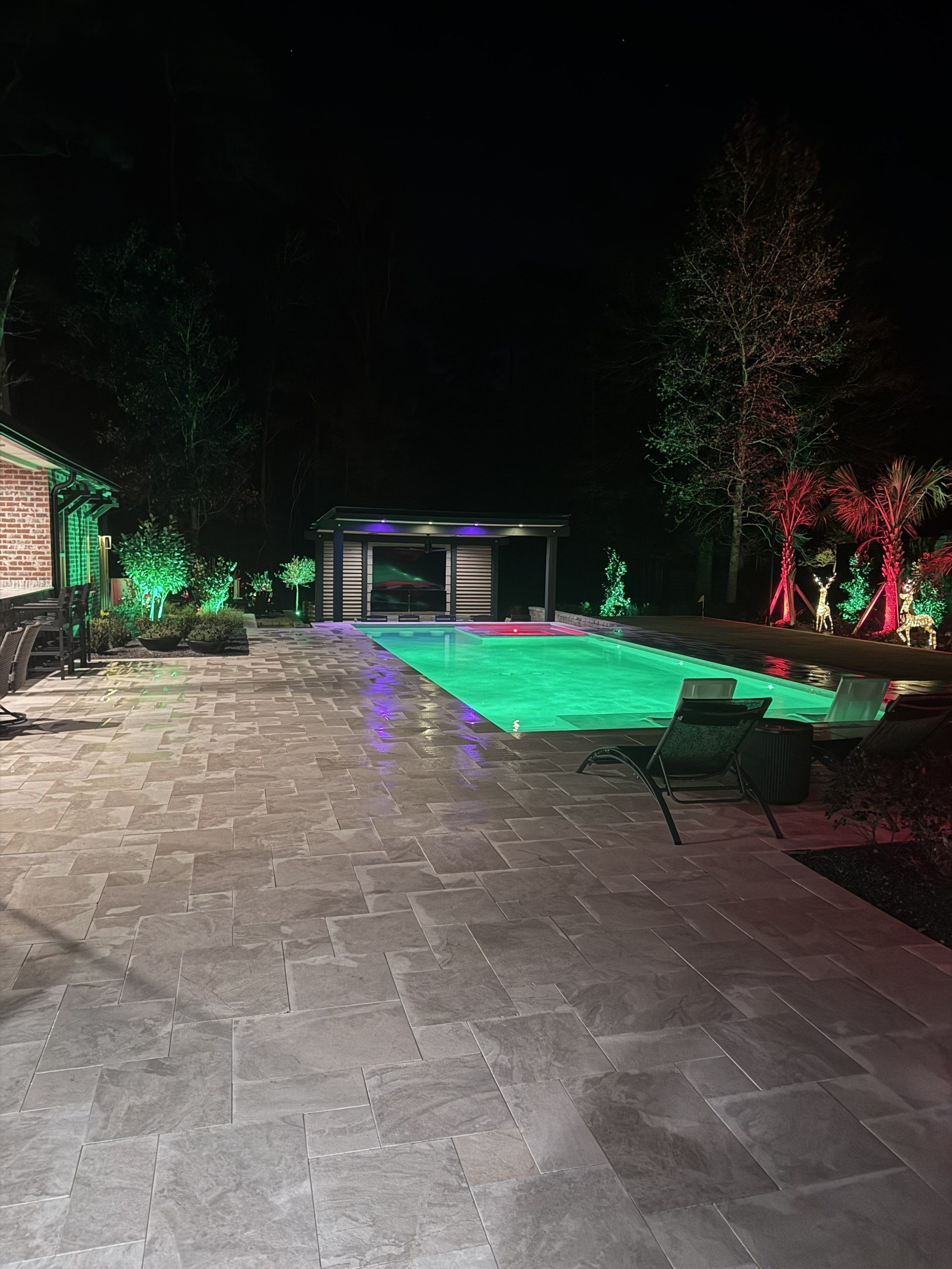 Nighttime patio with a lit green pool, lounge chairs, and trees beside a house